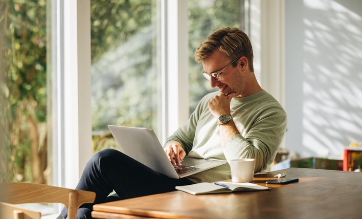 A man with blonde hair is sitting by a table researching Roth IRAs on his laptop computer.
